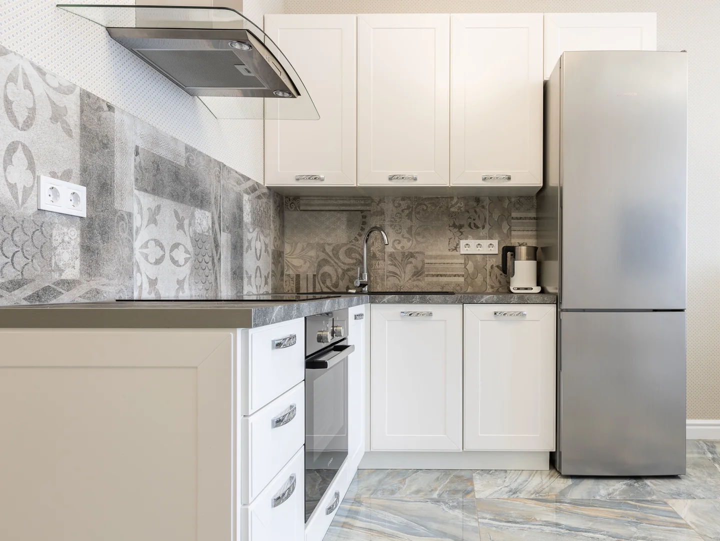 Kitchen with a silver-colored fridge, an oven between white cabinets and a kettle on the worktop.